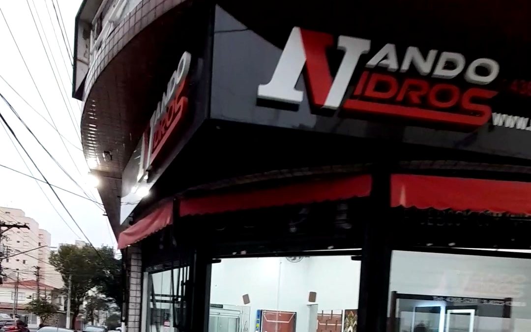 Fachada de esquina da loja Nando Vidros, com letreiro, toldo vermelho e vitrines de vidro